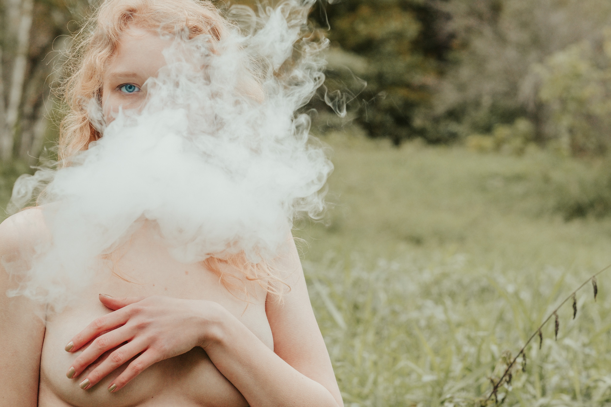 Blonde woman blowing smoke at camera while naked outside near trees